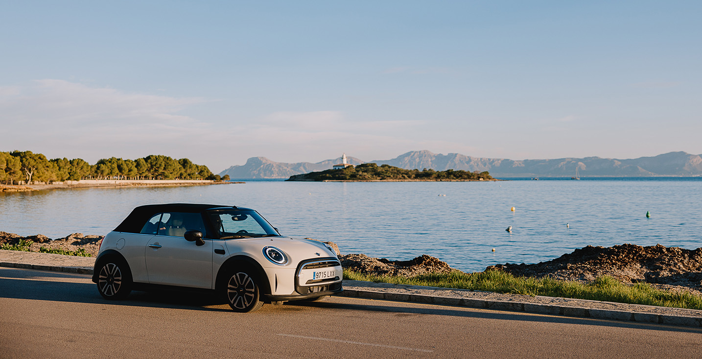 A white convertible Mini car is parked by a calm seaside road at sunset.