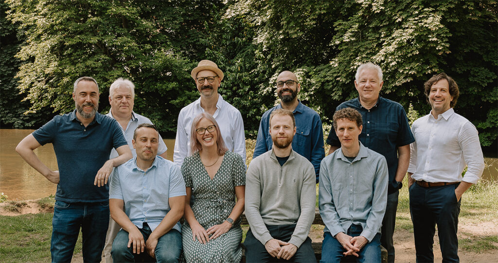 Group of ten people smiling posing outdoors on grass with trees in the background.