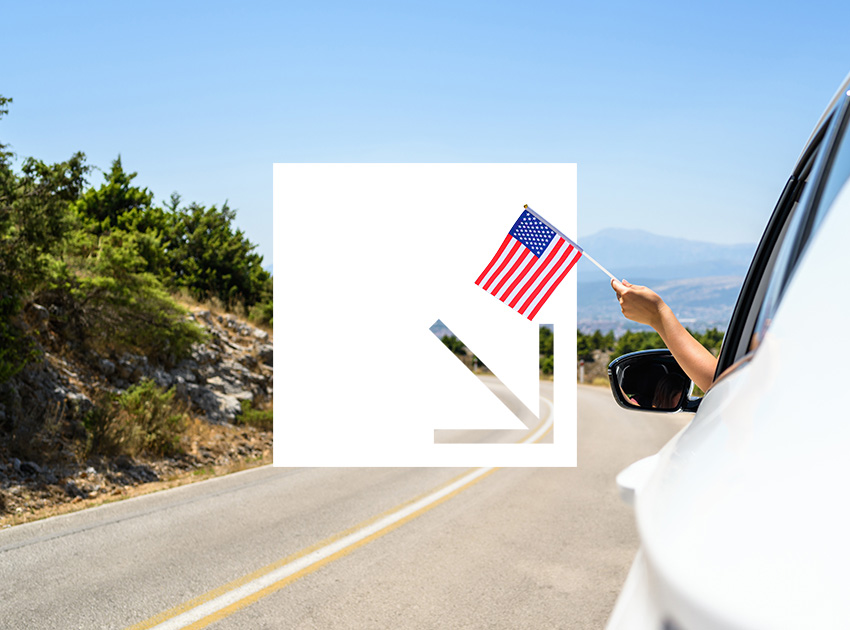 Hand waving a US flag from a car window on an open highway, highlighting the need for car hire excess insurance when driving in America.